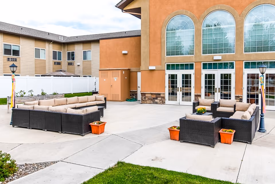 Outdoor patio area at Ellensburg Senior Living with two sets of black wicker outdoor sofas with beige cushions arranged around small tables. The patio is paved with concrete and decorated with orange flower pots and two lamp posts with colorful ribbons. The building in the background has large arched windows and multiple glass doors.