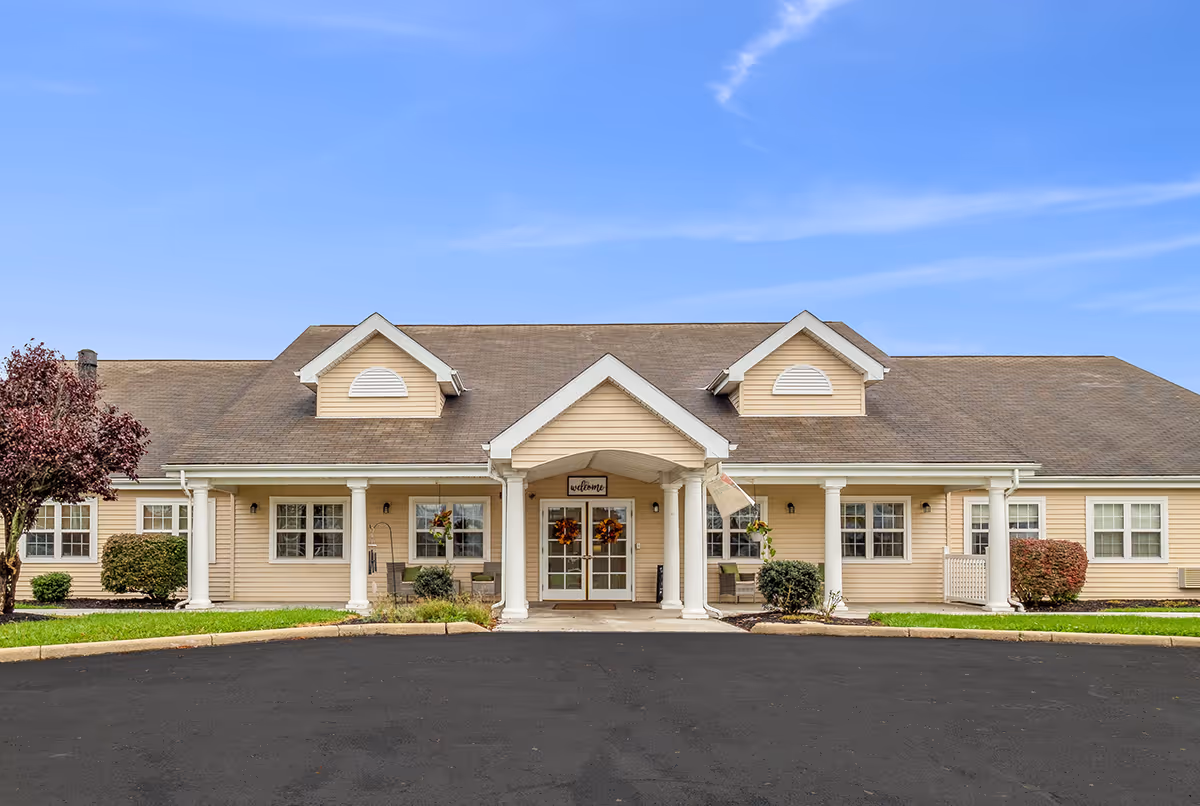 Front exterior view of a single-story senior living facility building with beige siding, white columns, and a covered entrance. The entrance has double glass doors decorated with wreaths and a small sign above that reads 'welcome'. There are shrubs and small trees on either side of the building, and the sky is clear and blue.