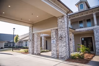 Entrance area of The Claiborne at Hattiesburg Assisted Living facility showing a covered driveway supported by brick columns, part of the building with balconies, and landscaped surroundings under a clear sky.