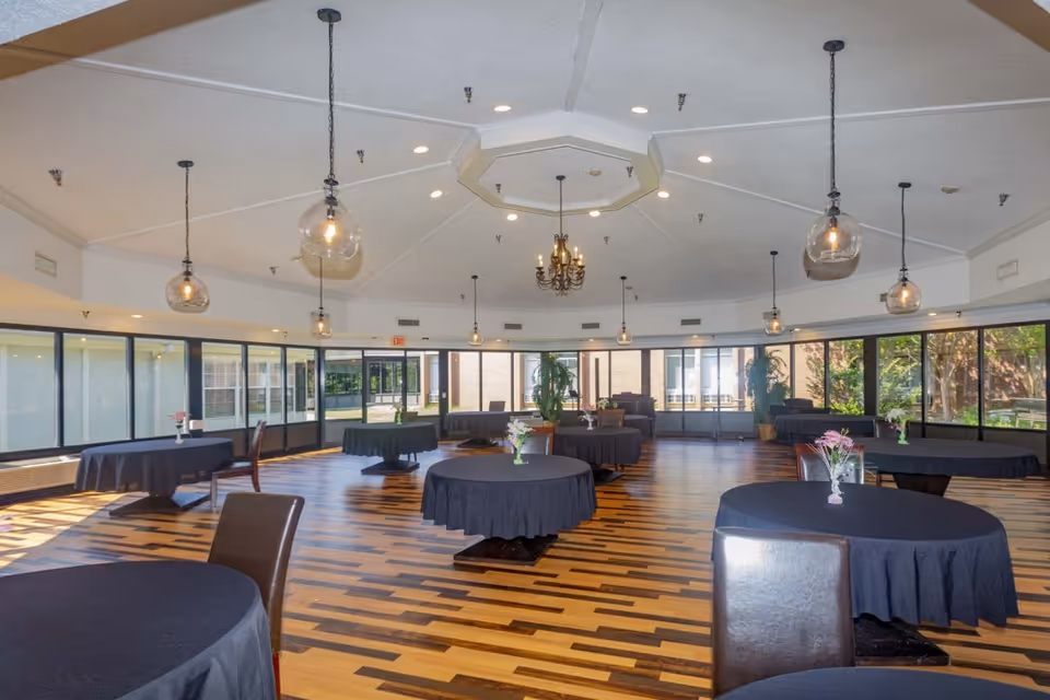 A spacious dining room with multiple round tables covered in black tablecloths, each adorned with small flower arrangements. The room features large windows allowing natural light to fill the space, modern pendant lights hanging from the ceiling, and a patterned wooden floor.