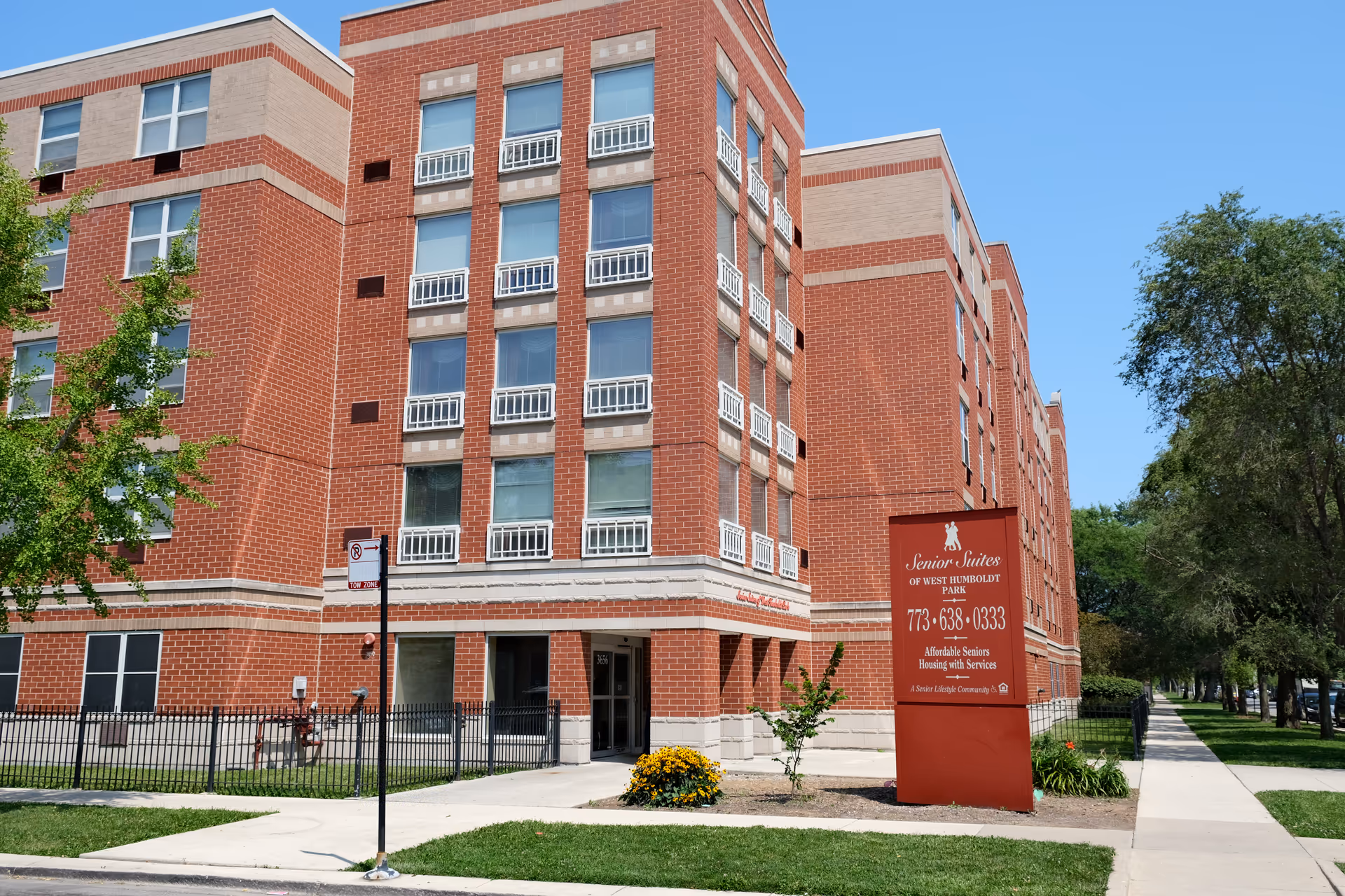 Exterior view of a multi-story brick building with multiple windows and a red sign in front that reads 'Senior Suites of West Humboldt Park, 773-638-0333, Affordable Seniors Housing with Services'. There is a sidewalk and green grass with trees around the building under a clear blue sky.