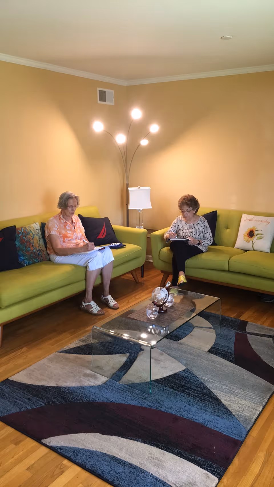 Two elderly women sitting on green sofas in a living room with yellow walls. They are both writing or reading on clipboards. The room has a modern glass coffee table on a multicolored rug, a floor lamp with multiple lights, and decorative pillows on the sofas.