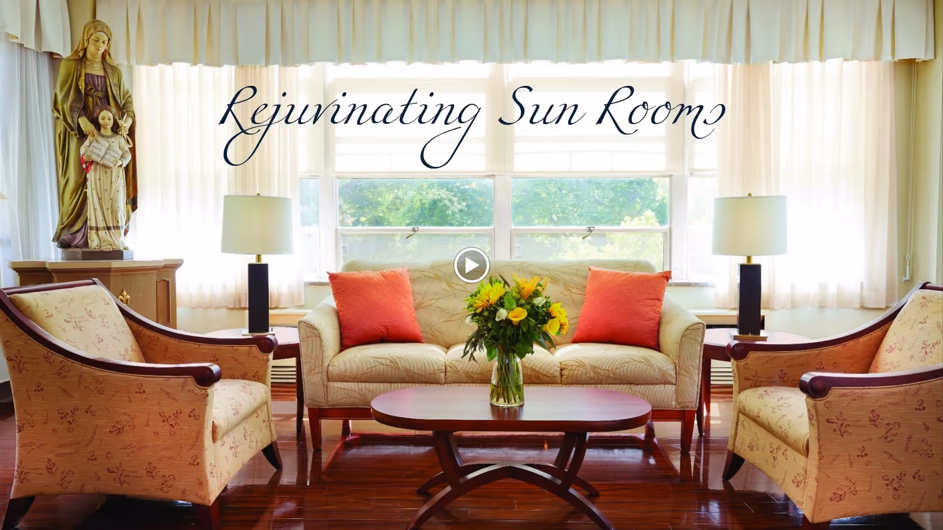 A bright sunroom with large windows covered by sheer white curtains, featuring a beige sofa with two coral-colored pillows, two matching armchairs with floral upholstery, a wooden coffee table with a vase of yellow flowers, two table lamps on side tables, and a statue of a woman holding a child in the corner.