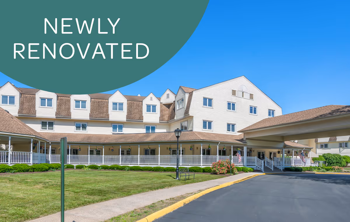 Exterior view of a large, newly renovated senior living facility with multiple windows, a covered entrance, and a wrap-around porch. The building is surrounded by a well-maintained lawn and a paved driveway under a clear blue sky.