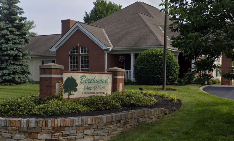 Exterior view of Birchwood Care Center, a single-story building with a pitched roof, surrounded by green grass, bushes, and trees. A stone and brick sign in front displays the facility name and logo.