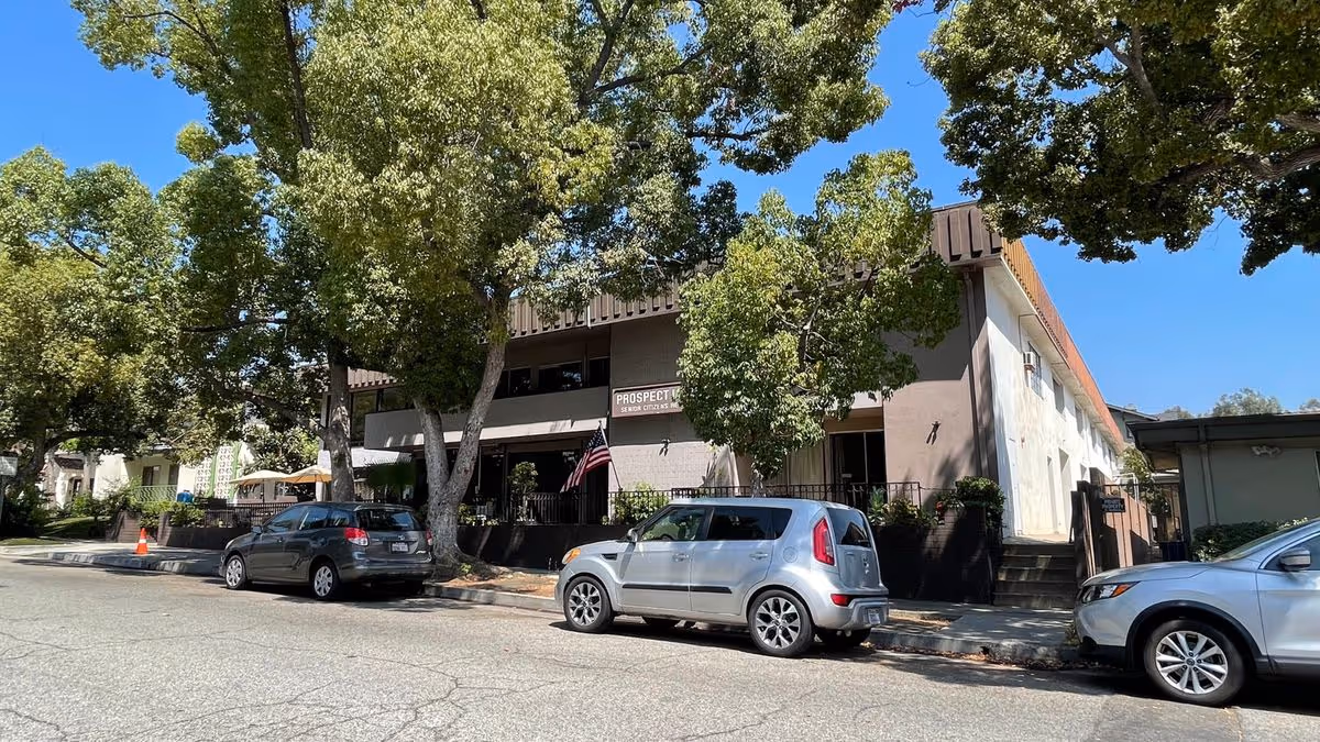 Street view of the Prospect Manor Assisted Living building with parked cars and large trees along the curb.