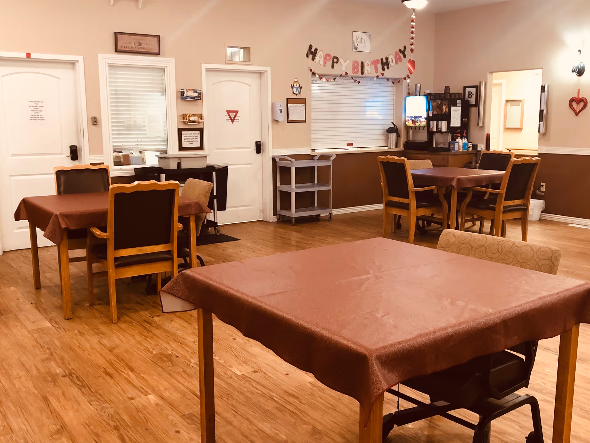 Communal dining room with several tables and chairs, a "Happy Birthday" banner, and a beverage station.
