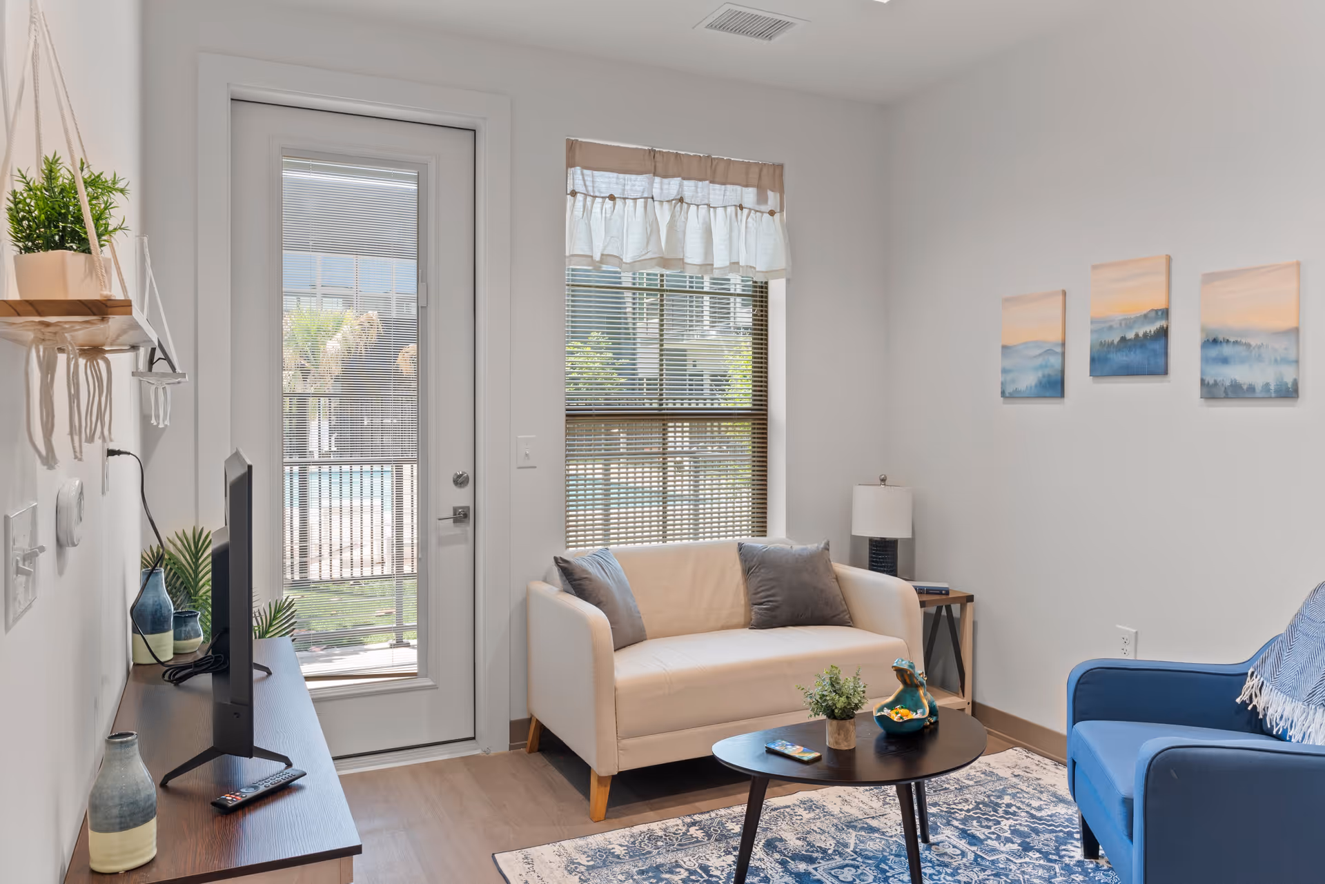 A bright and cozy living room with a white loveseat and a blue armchair. There is a small round black coffee table with decorative items on it, a patterned blue and white rug, and a wooden side table with a lamp. A TV sits on a dark wooden stand to the left. The room has a door with a glass panel and a window with blinds and a valance, letting in natural light. Three landscape paintings hang on the wall above the loveseat.