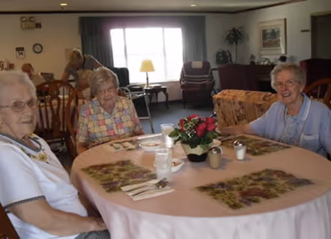 Three elderly women seated around a round dining table with place settings and a floral centerpiece in a communal dining/lounge area.