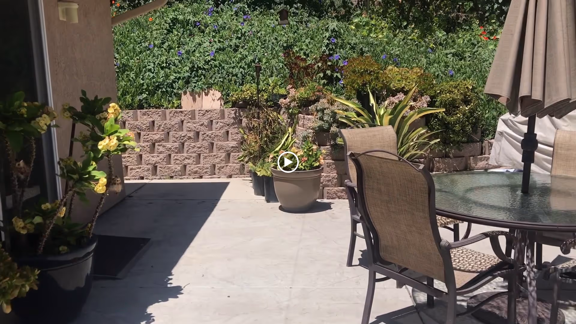 Outdoor patio area with a glass-top table and four chairs, an umbrella, potted plants, and a retaining wall with greenery and flowers in the background.