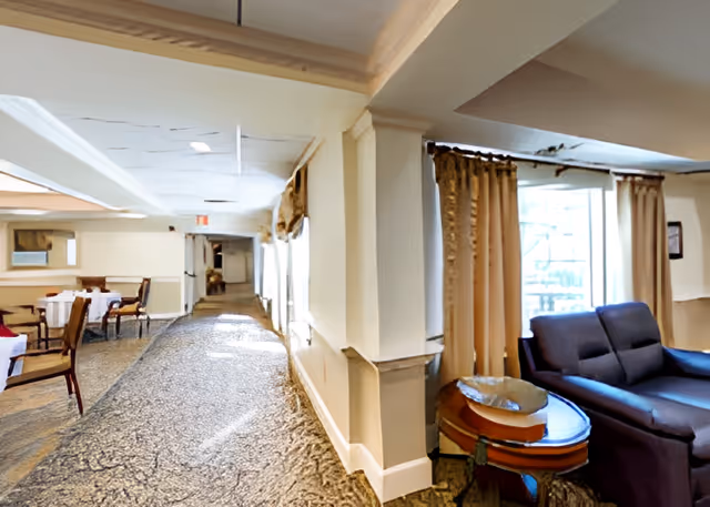 Carpeted interior corridor of a senior living facility with dining tables on the left and a seating area with a sofa on the right.