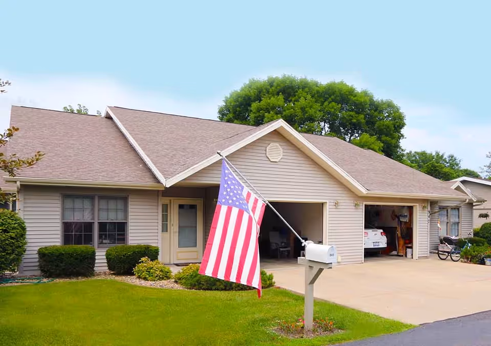 Single-story residential building with beige siding and a brown shingled roof, featuring a front door, two garage doors with one open showing a white car inside, a mailbox with the number 843, an American flag mounted on the mailbox, neatly trimmed bushes, and a green lawn under a clear blue sky.