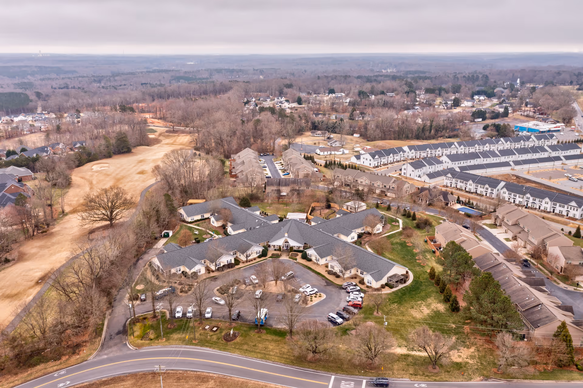 Aerial view of TerraBella Cramer Mountain senior living facility surrounded by trees, residential houses, and a golf course. The facility has a large, single-story building with a parking lot in front and landscaped grounds.