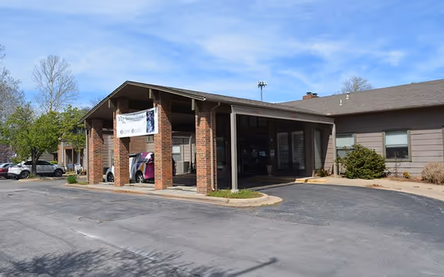 Exterior view of The Broadmoor Retirement Community building entrance with a covered drop-off area supported by brick pillars, a banner hanging from the roof, parked cars, and trees in the background under a clear blue sky.