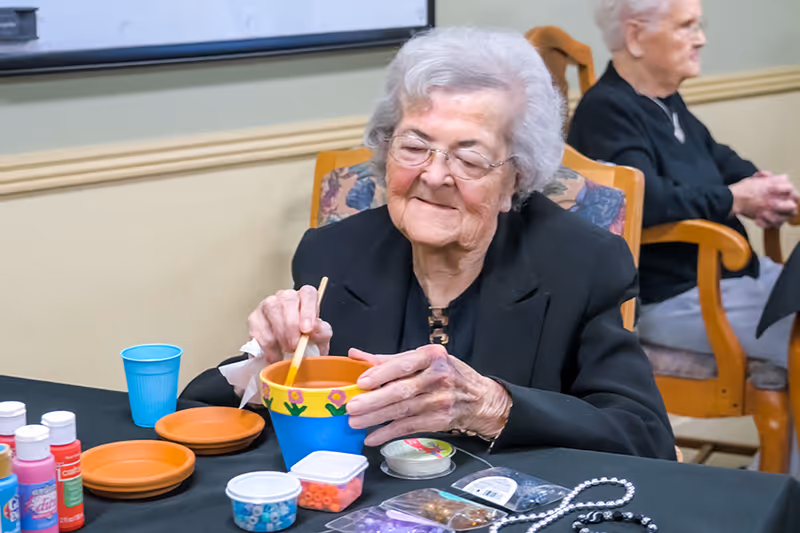 An elderly woman with white hair and glasses is sitting at a table, painting a colorful flower pot. Various craft supplies, including paint bottles, beads, and jewelry-making materials, are spread out on the table. Another elderly woman is seated in the background.