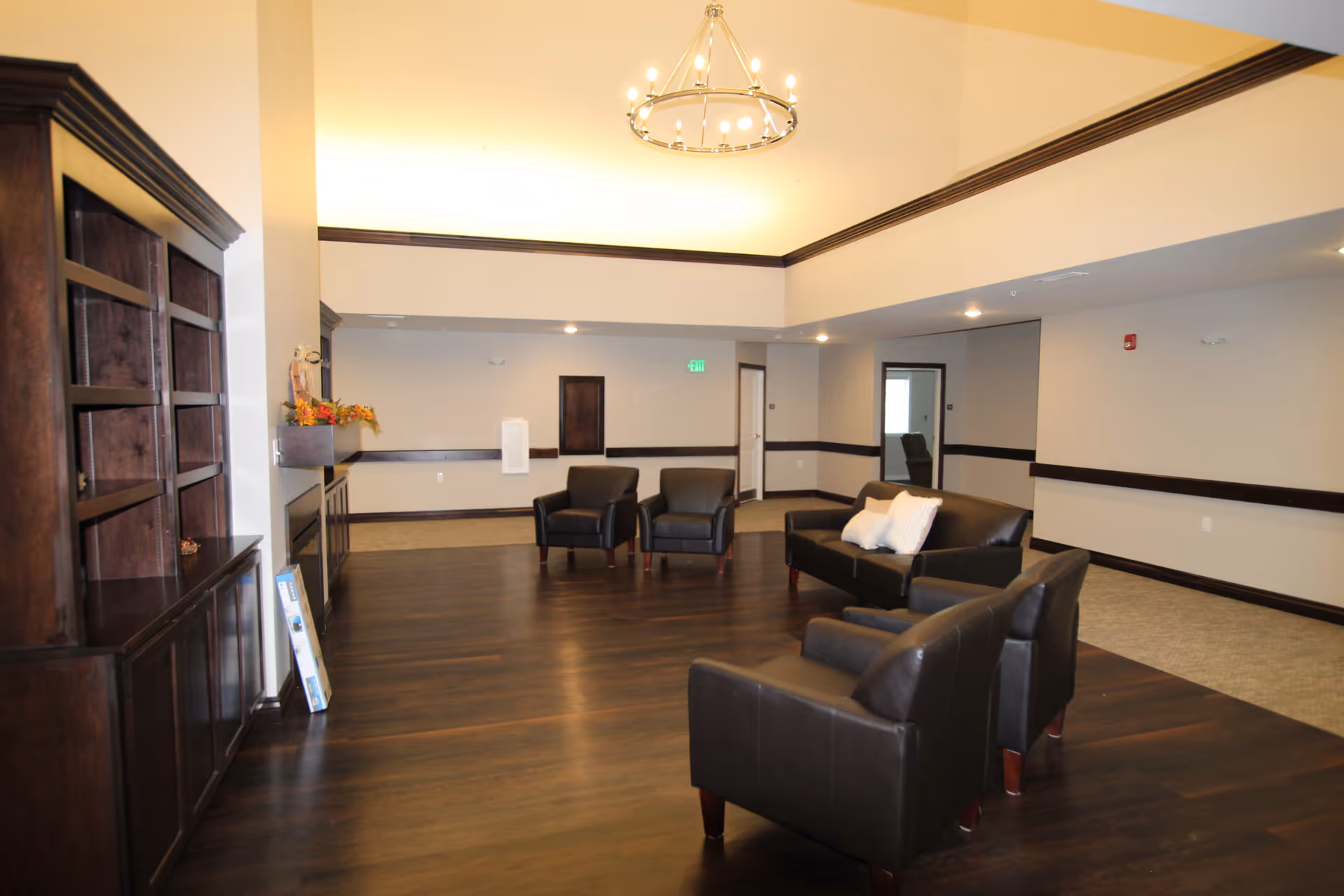 A spacious living room area with dark wood flooring and beige walls. The room features a dark brown leather sofa with white pillows, two matching armchairs, and a dark wood cabinet on the left side. A modern chandelier hangs from the ceiling, and there are several doorways and an exit sign visible in the background.