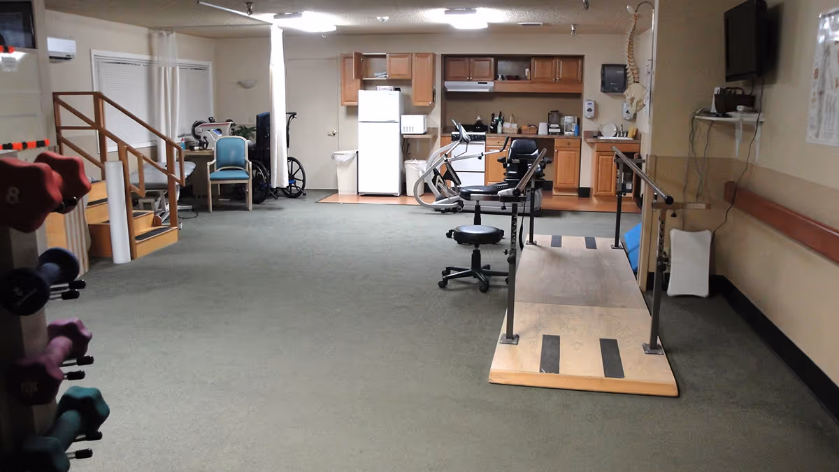 A rehabilitation center room with exercise equipment including a set of dumbbells, a parallel bar walking platform, and a stationary exercise bike. The room has a kitchenette area with cabinets, a refrigerator, and a microwave. There are chairs and a wheelchair near the back wall, and the floor is carpeted.