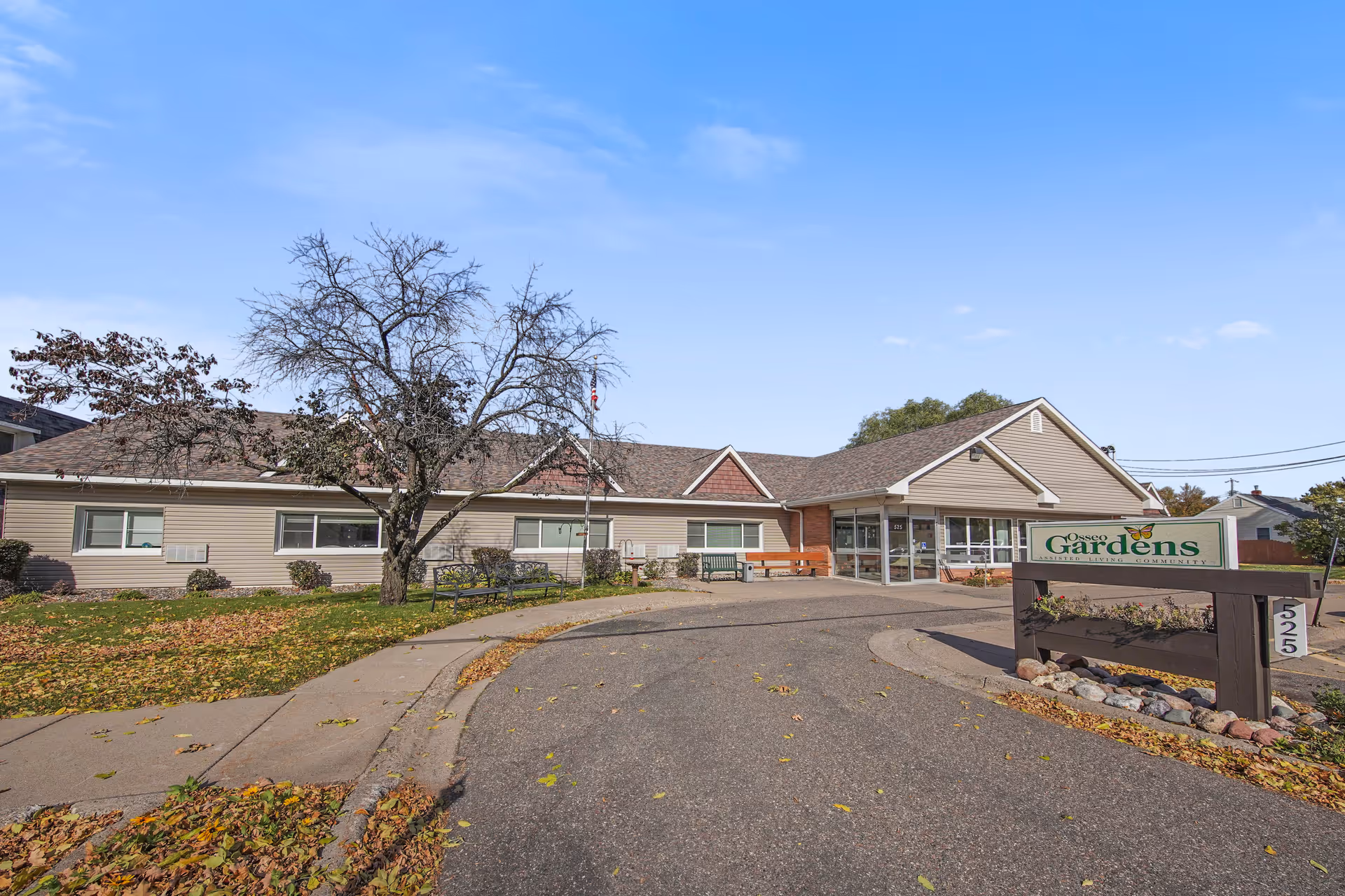 Exterior view of Osseo Gardens Assisted Living facility showing a single-story building with beige siding and a peaked roof. There is a curved driveway leading to the entrance, a leafless tree, benches, and a sign with the facility name and address number 525. The sky is clear and blue.