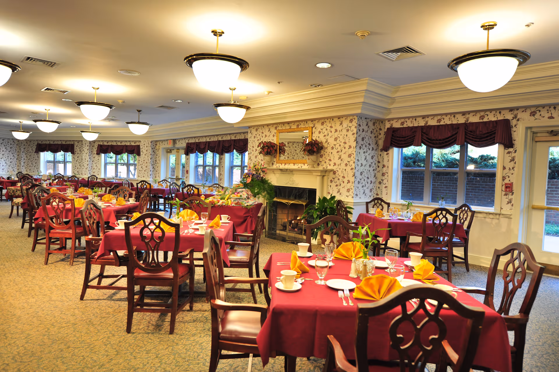 A spacious dining room with multiple tables covered in red tablecloths, each set with yellow folded napkins, white plates, cups, and glassware. The room features wooden chairs, floral wallpaper, large windows with maroon valances, and a fireplace with a mirror and floral decorations above it. Ceiling lights illuminate the room warmly.
