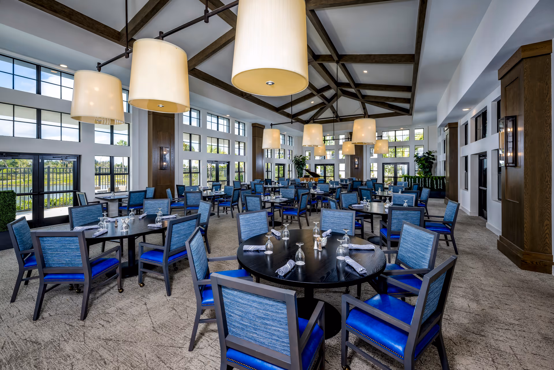Large bright dining room with round dark wood tables and blue upholstered chairs beneath oversized pendant lights and high beamed ceilings, with many windows.