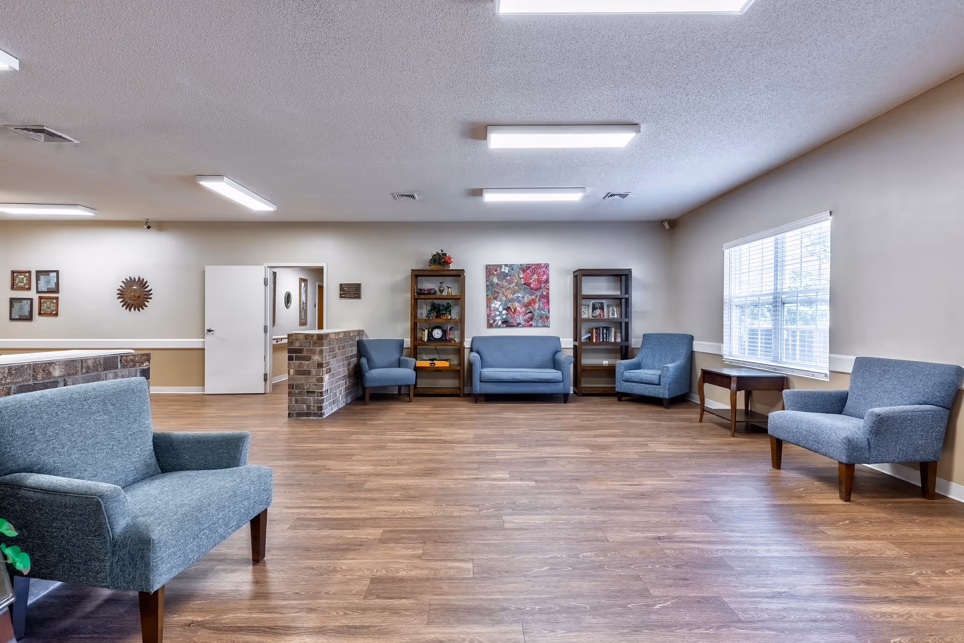 A spacious living room area with wooden flooring and beige walls. The room features several blue upholstered chairs and a loveseat arranged around the perimeter. There are two wooden bookshelves with decorative items and books, a floral painting on the wall, a window with white blinds letting in natural light, and a small wooden side table. The ceiling has multiple rectangular fluorescent light fixtures.