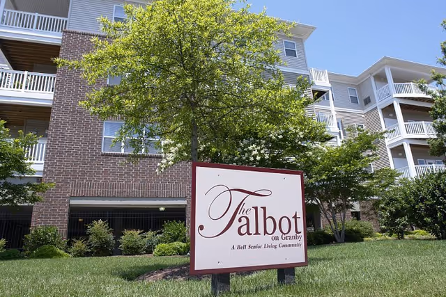 Exterior view of a multi-story senior living community building with balconies, surrounded by green trees and grass. In the foreground, there is a sign that reads 'The Talbot on Granby, A Bell Senior Living Community.'