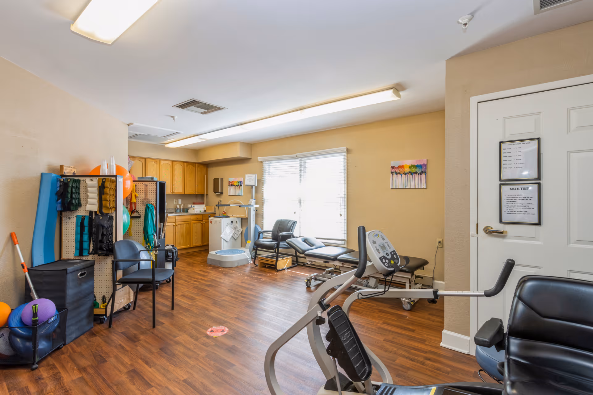 A physical therapy or rehabilitation room with exercise equipment, therapy balls, chairs, a treatment table, and wooden cabinets along the back wall. The room has wood flooring, beige walls, and a window letting in natural light.