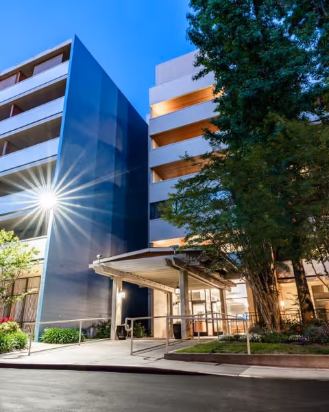 Exterior view of a multi-story senior living facility building at dusk with illuminated entrance, large trees, and landscaped greenery.