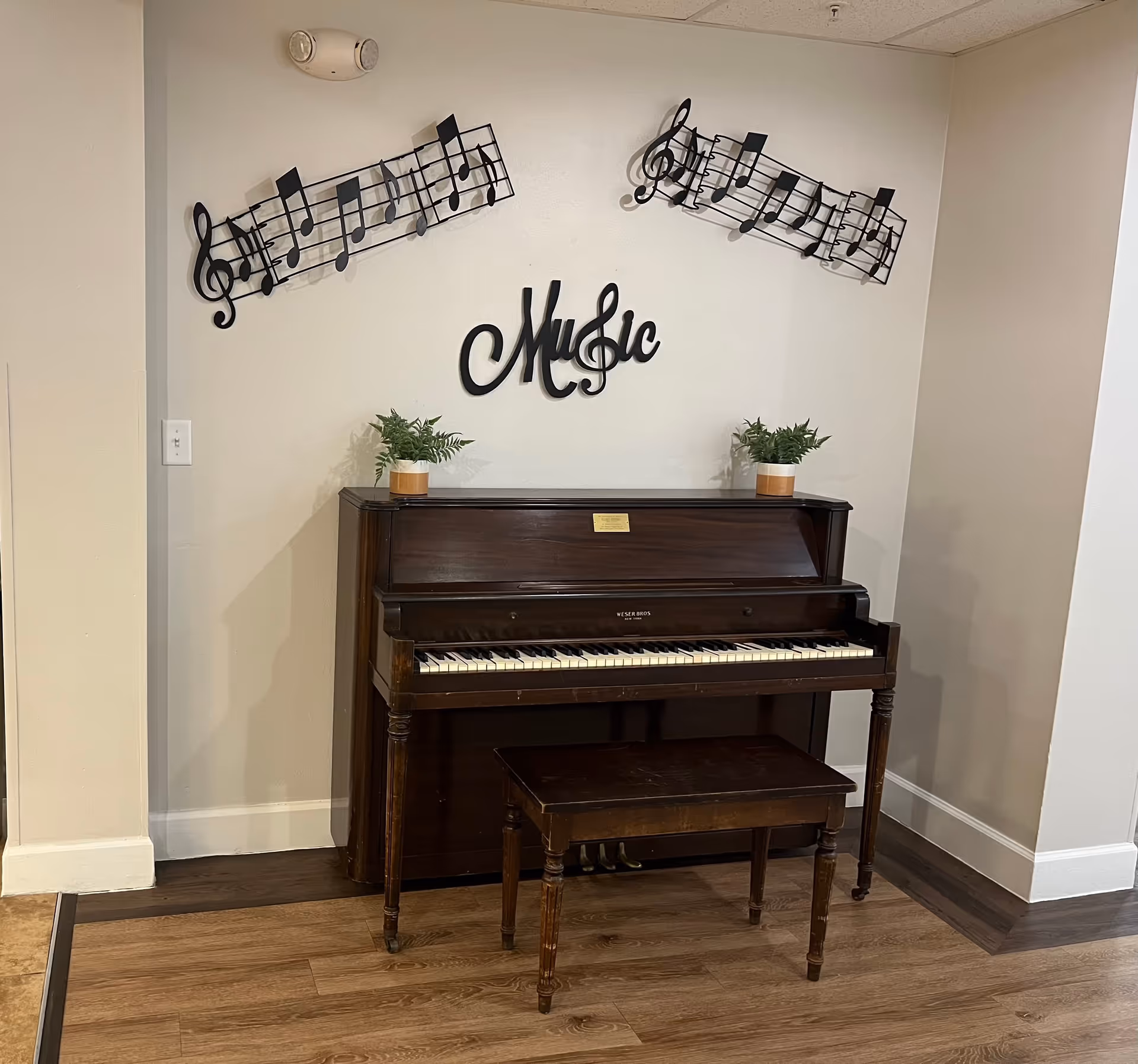 An upright wooden piano with a matching bench is placed against a beige wall. Above the piano, there are two decorative metal musical notes and the word 'Music' in black metal art. Two small potted plants sit on top of the piano. The floor is wood with a small section of tile visible on the left.