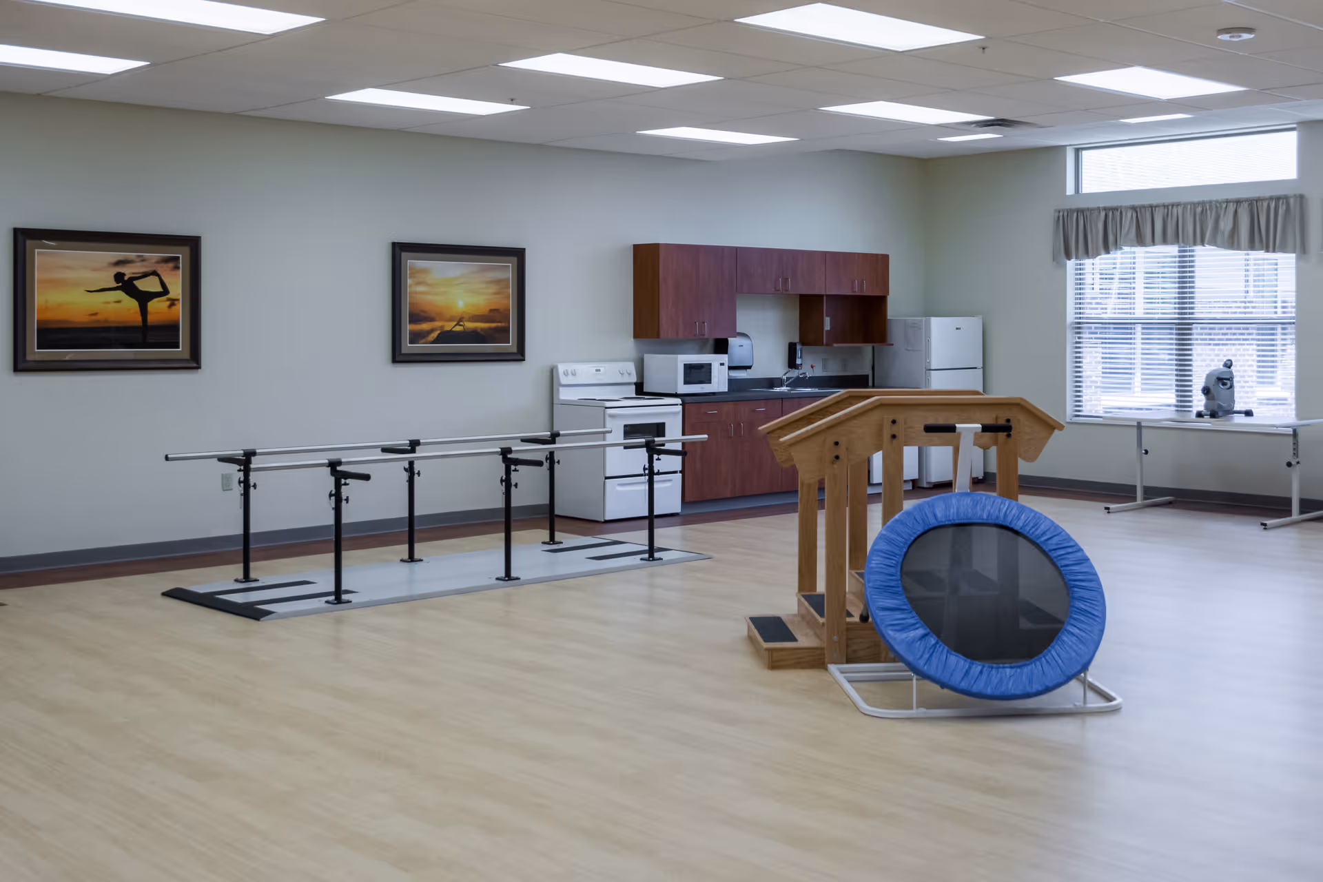 A spacious room with light wood flooring and pale green walls featuring parallel bars for walking exercises, a small trampoline, and wooden handrails for physical therapy. The back wall has a kitchenette with a stove, microwave, refrigerator, and cabinets. Two framed pictures of sunset scenes hang on the wall. A large window with blinds and a valance lets in natural light, and a small exercise bike is positioned near the window.