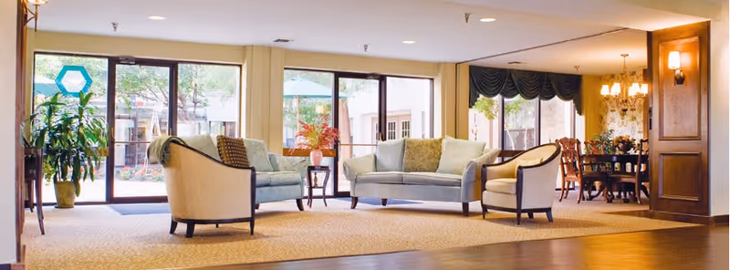 A bright and spacious living room area with large windows and glass doors looking out to an outdoor patio. The room features a light blue sofa, two armchairs, a small side table with a vase of flowers, and a dining area with a chandelier in the background. There are plants near the windows and warm lighting throughout.