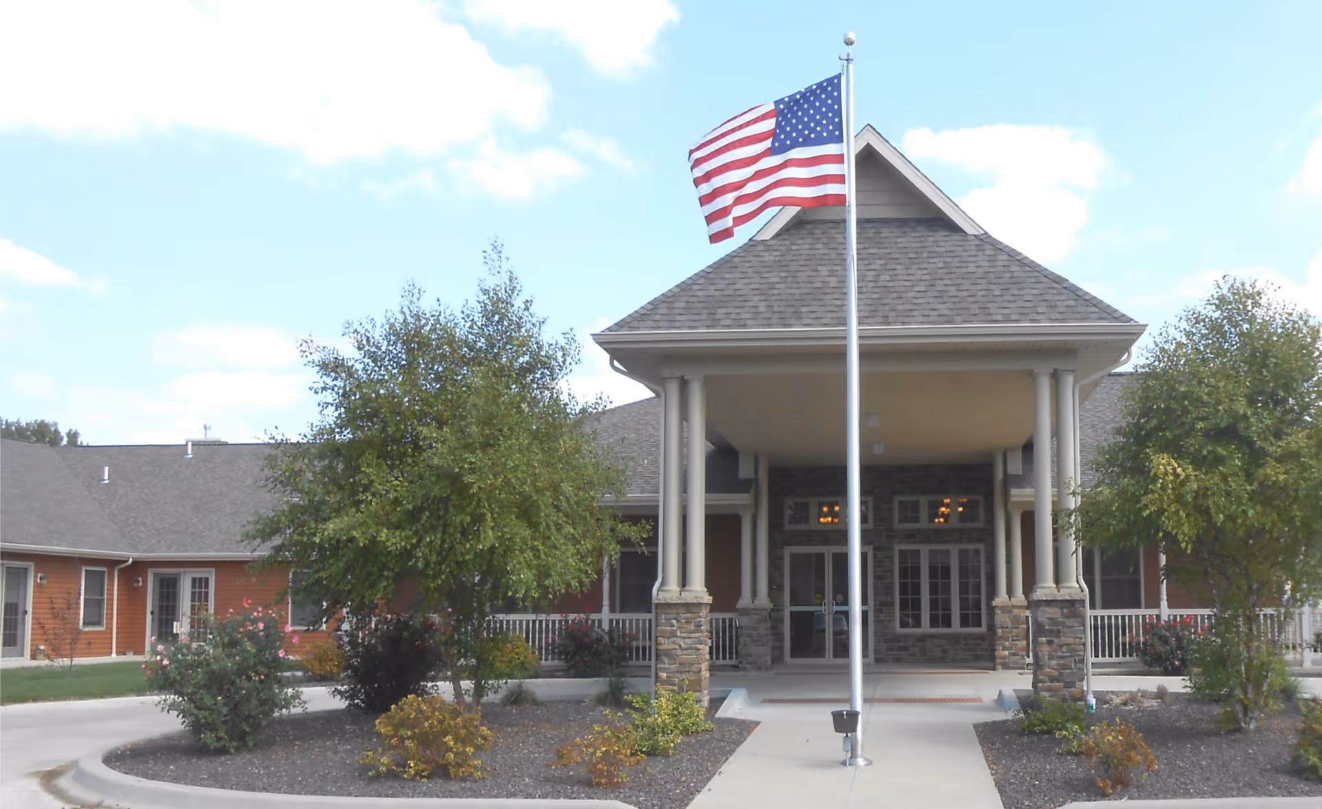Front entrance of a senior living building with a covered porte-cochère, an American flag on a flagpole, and landscaped beds.