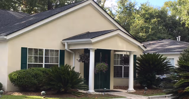 Exterior view of a single-story building with beige walls, green shutters, and a dark roof. The entrance features a small porch with white columns and hanging flower baskets. There are bushes and landscaped plants around the building, and trees in the background.