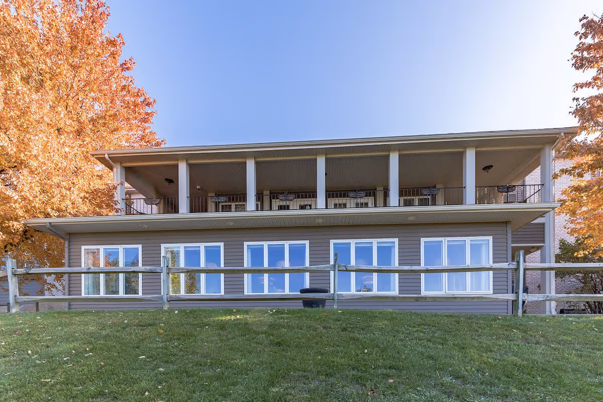 Exterior view of a two-story building with a covered balcony on the upper floor and multiple windows on the lower floor. The building is surrounded by green grass and autumn-colored trees under a clear blue sky.