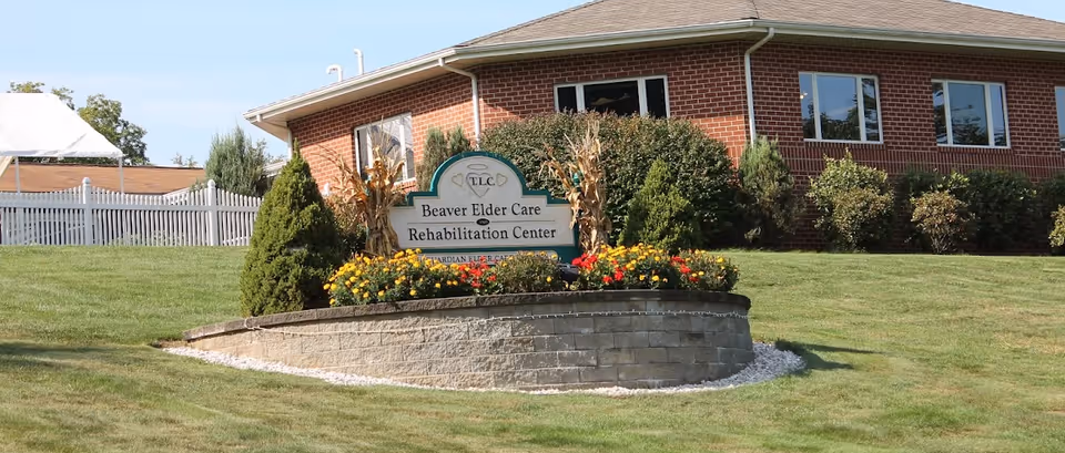 Exterior view of a brick building with a landscaped garden in front featuring a sign that reads 'Beaver Elder Care Rehabilitation Center'. The garden has colorful flowers and trimmed bushes, and there is a white fence in the background under a clear blue sky.