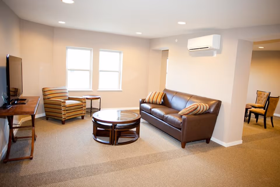 A bright living room with beige walls and carpeted floor featuring a brown leather sofa with two patterned pillows, a striped armchair, a round glass coffee table, and a flat-screen TV on a wooden stand. There are two windows letting in natural light and an air conditioning unit mounted on the wall. In the background, there are two upholstered chairs near an open doorway.