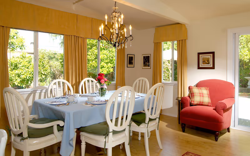 Sunlit dining room with a set table covered by a blue tablecloth, white chairs, a chandelier, and a red armchair by a sliding glass door.