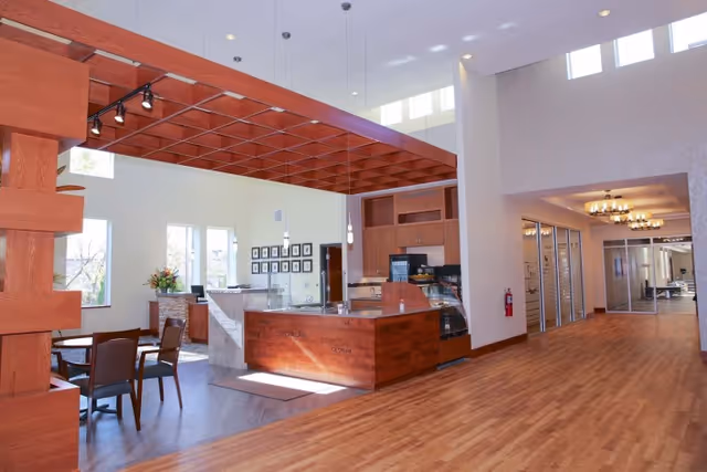 Interior view of a senior living facility showing a spacious area with wooden flooring and a wooden reception or service counter with a lattice wooden ceiling feature above it. There are tables and chairs to the left near large windows letting in natural light. The hallway to the right has glass doors and modern light fixtures on the ceiling.