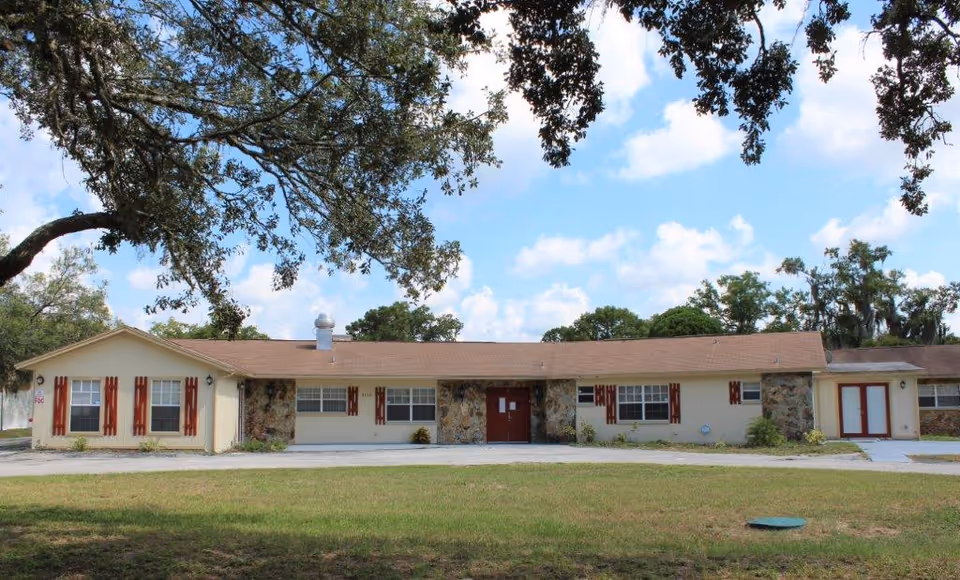 Single-story beige ranch-style building with stone accents, red shutters and doors, a front lawn and a large tree under a blue sky.