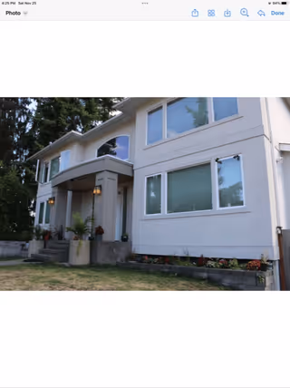 Exterior front view of a two-story residential building with large windows, a covered entrance with two columns, outdoor lighting fixtures, and a small garden bed with plants and flowers along the front.
