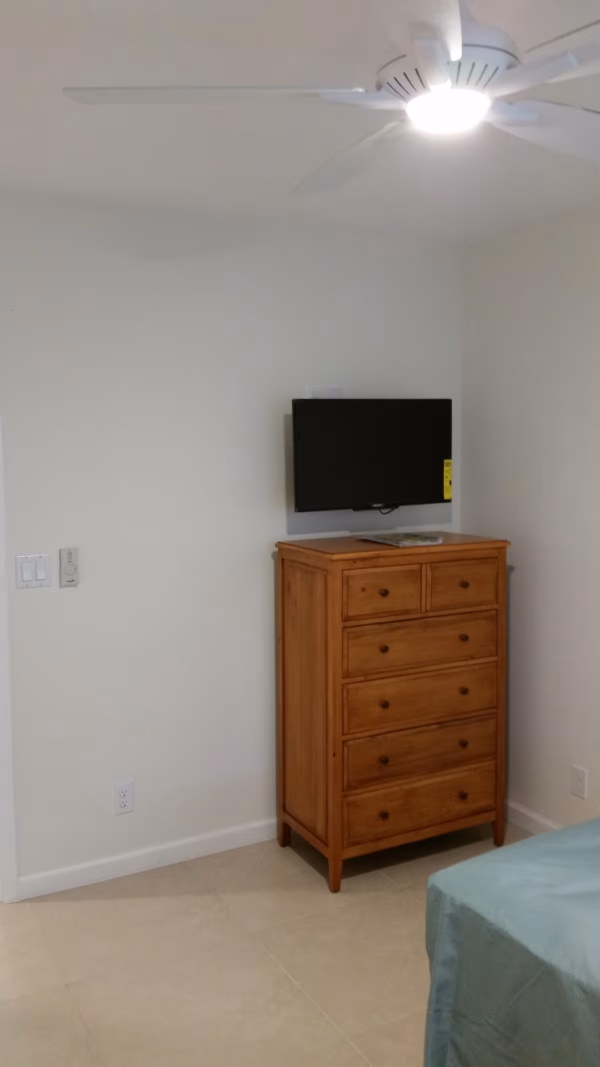 A corner of a bedroom with a wooden chest of drawers, a flat-screen TV mounted on the wall above it, a ceiling fan with a light, and part of a bed with a blue bedspread visible in the foreground.