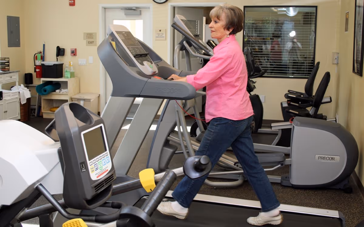 An elderly woman in a pink shirt and jeans is walking on a treadmill in a fitness room equipped with various exercise machines including treadmills and stationary bikes.