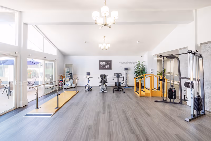 Bright and spacious rehabilitation gym with exercise equipment including stationary bikes, parallel bars for walking practice, a wooden balance bridge, and a cable machine. Large windows on the left side let in natural light and provide a view of an outdoor patio area with tables and umbrellas. The walls are white with motivational signs, and the floor is covered with gray wood-like flooring.