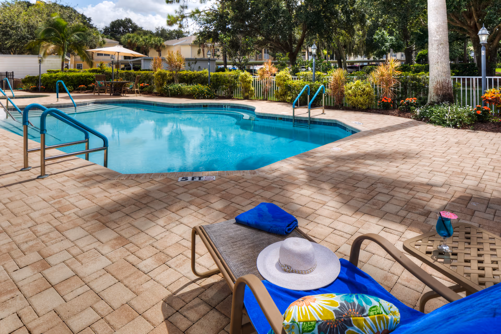 Outdoor swimming pool area with clear blue water, surrounded by a brick-paved deck. There are metal handrails for pool entry, a lounge chair with a colorful pillow, a blue towel, and a white sun hat. A small table next to the chair holds a blue drink with a pink umbrella. In the background, there are green bushes, trees, and residential buildings under a partly cloudy sky.