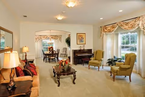 A bright and spacious living room with beige carpet and walls, featuring a tan sofa with red cushions, two green armchairs, a wooden coffee table with a floral centerpiece, a piano against the far wall, and large windows with floral valances allowing natural light to fill the room. In the background, a dining area with a round table and chairs is visible through an open archway.