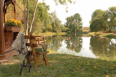 A peaceful outdoor scene featuring a small wooden bench and a fishing rod holder with fishing rods next to a wooden structure adorned with a pot of yellow flowers. In the background, there is a calm pond surrounded by green trees and grass.