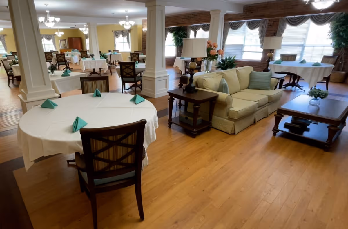 A spacious dining area with multiple round tables covered with white tablecloths and green folded napkins. The room features wooden floors, several chairs around each table, and a cozy seating area with a beige sofa, side tables with lamps, and a coffee table. Large windows with drapes allow natural light to fill the room, and decorative plants add a touch of greenery.