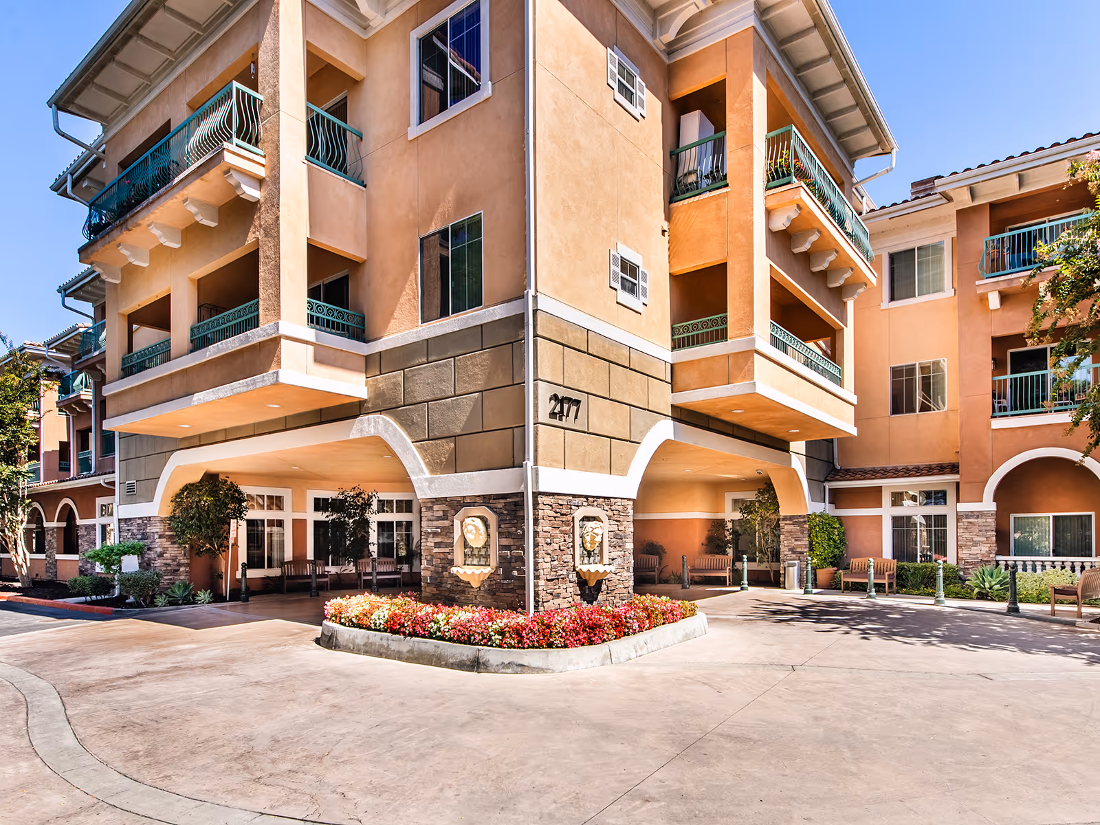 Front entrance and facade of a multi-story stucco building with balconies, arched drive-through, and a flower bed.