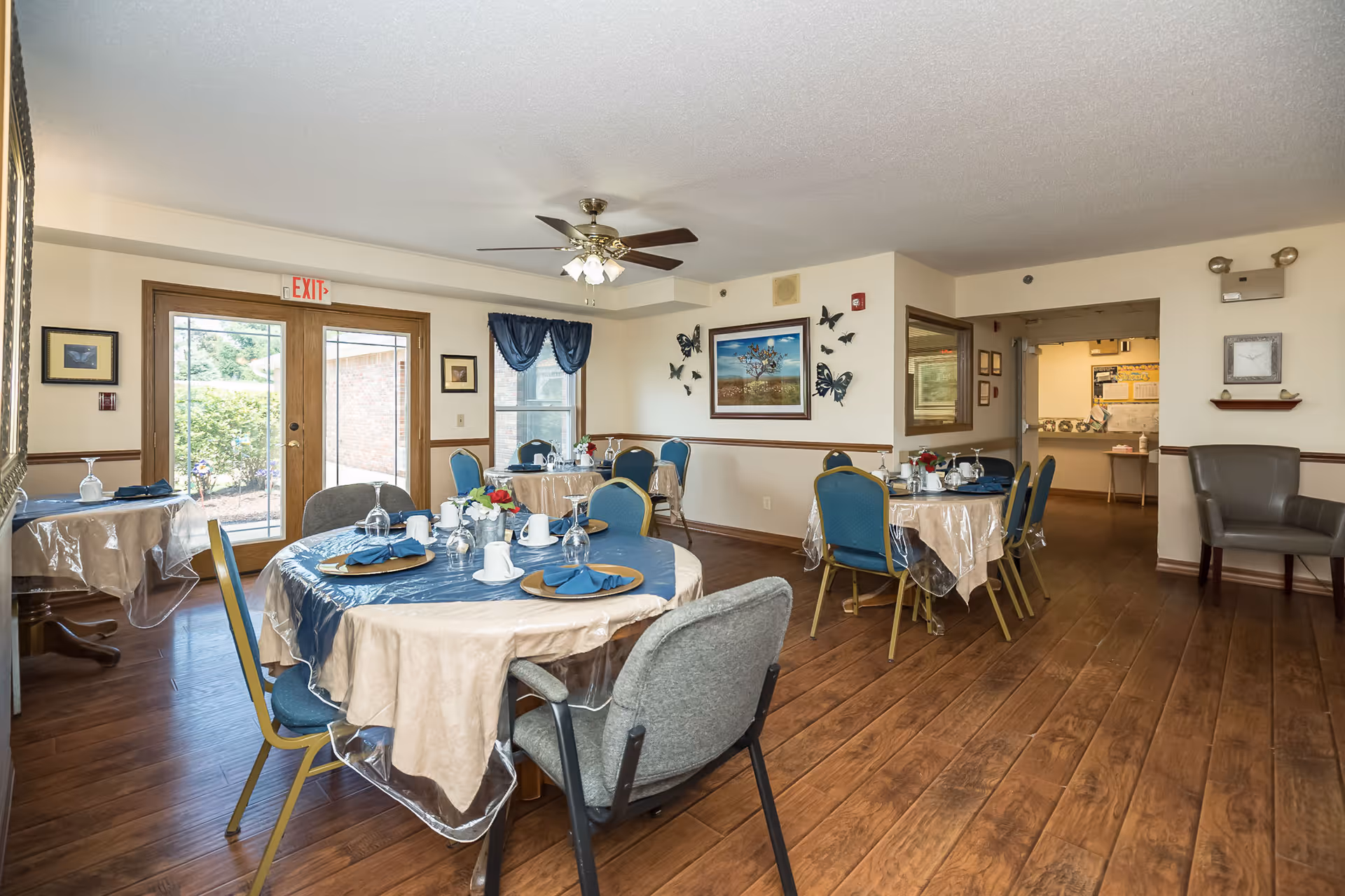 A dining room in a senior living facility with several round tables covered with beige and blue tablecloths, set with plates, cups, and napkins. The room has wooden flooring, a ceiling fan with lights, blue curtains on the window, and wall decorations including framed pictures and butterfly wall art. There are multiple chairs around the tables and an exit door leading outside.
