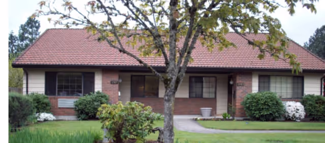 Single-story brick building with a red tiled roof, two large windows, and a central entrance door. A tree with green leaves stands in the front yard, which has a well-maintained lawn and bushes on either side of the walkway leading to the door.